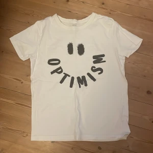 Vit t-shirt från Name It - En vit t-shirt från Name It med texten 'OPTIMISM' tryckt på framsidan i svart. T-shirten är kortärmad och tillverkad av ekologisk bomull.