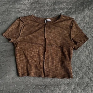 Brun ribbad topp från Divided - En brun ribbad topp från HM Divided med korta ärmar och en rund halsringning. Toppen har en synlig söm som löper horisontellt över bröstet och vertikalt längs mitten. Använd få gånger 
