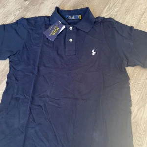 Mörkblå pikétröja från Polo Ralph Lauren - En mörkblå pikétröja från Polo Ralph Lauren med en broderad logotyp på bröstet. Tröjan har en klassisk krage och två knappar vid halsen. tagsen är ej kvar. tröjan är 10/10 skick
