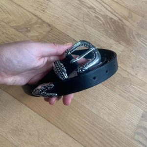 Svart bälte med silverdetalj från Gina Tricot  - Ett svart bälte med silverdetalj framtill från Gina Tricot. Bältet är i storlek M/L och är i bra skick. Det har även gjorts ett extra hål i bältet (kan man se på bild 3).