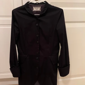 Svart långärmad klänning från Noa Noa - Säljer en elegant svart kappa från Noa Noa. Kappan har en klassisk design med knappar hela vägen ner och en fin krage. Den är perfekt för både vardag och fest. Materialet känns mjukt och bekvämt, och den har en normal passform. Kappan är i nyskick och redo att bäras av någon annan. Passar perfekt till höst och vinter!