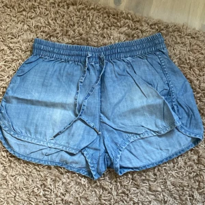 Blå jeansshorts från &Denim - Säljer ett par snygga blå jeansshorts från &Denim i storlek XS. De har en elastisk midja med knytband och två fickor både fram och bak. Perfekta för sommaren och riktigt bekväma att ha på sig. Passar både till vardags och till stranden!
