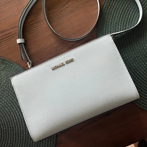 Vit axelväska från Michael Kors - Säljer en snygg ljusblå axelväska från Michael Kors. Väskan är i skinn och har en justerbar axelrem. Insidan är fodrad med Michael Kors-logotyp och har flera fack för enkel organisering. Väskan är i nyskick och redo för en ny ägare!