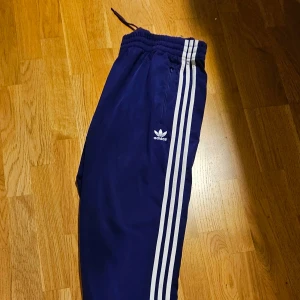 Säljes: Adidas träningsbyxor - Säljer ett par snygga och bekväma Adidas träningsbyxor. Byxorna är mörkblå med de klassiska tre vita ränderna längs sidorna samt Adidas logotypen på ena benet. De är i storlek XL och har en elastisk midja med dragsko för en perfekt passform. 