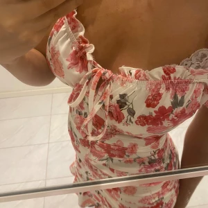 Blommig klänning med volangdetaljer - En vacker klänning med blommigt mönster i rosa och rött på en vit bakgrund. Klänningen har volangdetaljer vid halsringningen och nederkanten samt en knytning framtill. Den är ärmlös och har en tight passform.