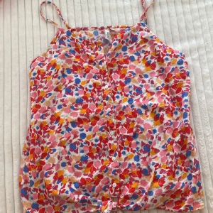 Blommig linnetopp från Little Pieces - En färgglad blommig linnetopp från Little Pieces med knytning framtill. Toppen har smala axelband och knappar längs framsidan. Mönstret består av blommor i rosa, rött, blått, gult och vitt.