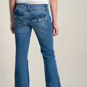 Blå bootcut jeans - Säljer ett par snygga blå bootcut jeans. De har en klassisk femficksdesign med två bakfickor som har knappar. Jeansen är i ett mjukt och bekvämt material, perfekt för vardagsbruk. De är lågmidjade och har en tight passform upptill som går ut i en bootcut nertill.