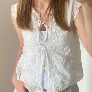 TOPP MED BRODERIER OCH KNYTNING🤍 - ORDINARIE PRIS 375 kr. Har aldrig användt den. Rund krage, ärmlös, broderier och kombinerad infälld spets i samma färg. Knäppning fram med knytning. Skriv ifall du har några funderingar ang plagget.😊