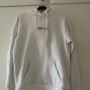 Adidas huvtröja - En vit hoodie från Adidas med en känguruficka och dragsko i huvan. Tröjan har Adidas-logotypen och texten 'The brand with the 3 stripes' på bröstet. Den har långa ärmar och är i gott skick.