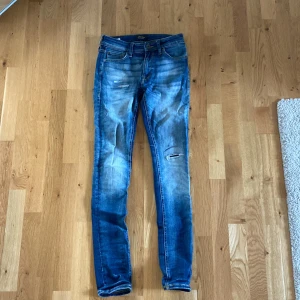 Blå jeans från Jack & Jones - Säljer ett par snygga blå jeans från Jack & Jones i storlek 28/32. De har en slim passform och är i bra skick med en cool slitning på ena benet. Perfekta för både vardag och fest!