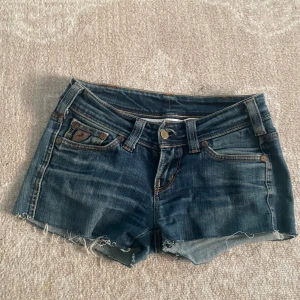 Mörkblå jeansshorts från Lois - Säljer ett par mörkblå jeansshorts från Lois i storlek 34. De är lågmidjade och har en snygg, sliten look med fransiga kanter. Perfekta för sommaren! Shortsen har fem fickor och knappar samt dragkedja framtill. De är i bra skick och redo för en ny ägare.