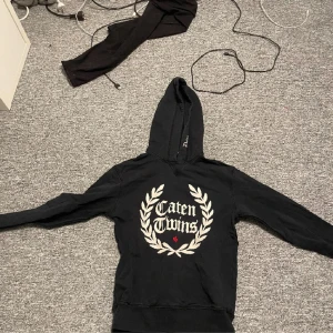 Svart hoodie från Dsquared2 - Säljer en snygg svart hoodie från Dsquared2 med texten 'Caten Twins' och en krans på framsidan. Den är i storlek 14Y och har en skön passform. Perfekt för höst och vinter! 🖤