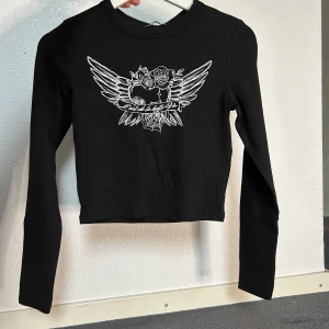 Svart croppad tröja från Bershka - Säljer en snygg svart croppad tröja från Bershka. Den har ett coolt vitt tryck med vingar och blommor på framsidan. Tröjan är långärmad och perfekt för både vardag och fest. Materialet är mjukt och bekvämt, vilket gör den skön att ha på sig hela dagen. Passar perfekt till höst- och vårsäsongen!