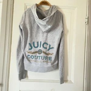 Grå hoodie från Juicy Couture - Säljer en superfin grå hoodie från Juicy Couture. Den har en dragkedja framtill och en stor luva. På baksidan finns det ett stort Juicy Couture-tryck med glittriga detaljer i blått och guld. några pärlor från trycker har lossnat men inget man tänker på. Perfekt för en avslappnad och stilren look! skriv egna prisförslag!💓