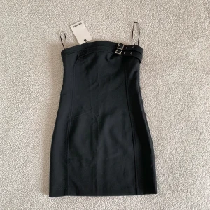 Pull and Bear strapless buckle dress / svart mini klänning - Otroligt snygg miniklänning från Pull and Bear med silvrigt spänne vid bysten. Ny med tags kvar, utan anmärkningar. Sitter som ett smäck och formar kroppen fint. Håller sig på plats då den har silikonband runt bysten.