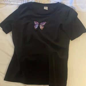 Svart sammet t-shirt med en fjäril  - Hej! Nu säljer jag min svarta sammet t-shirt med en lila fjäril på från Lindex.  Använd fåtal gånger. Storlek: 146/152. Nypris: ca 150kr. Mitt pris: 80kr. Köparen betalar frakten. 