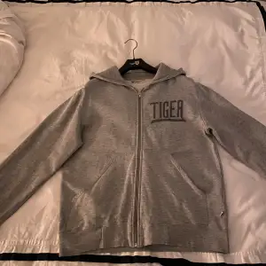 Grå Zip hoodie från Tiger of Sweden med broder Tiger på vänstra bröstet. Bra skick, storlek Medium. Vid vidare funderingar är det bara att höra av sig!