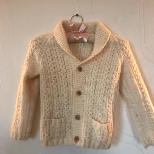 Stickad kofta - Gräddvit stickad kofta som är lite tjockare. Den knäpps med knappar och så har den två fickor. Den är stickad i väldigt fint mönster och har en krage vid nacken. Det står ingen storlek, skulle säga att den passar ungefär storleken 128. Väldigt bra skick. 