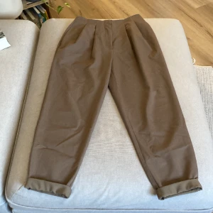 Kostymbyxor nakd - Bruna kostymbyxor från nakd med veckas midja. Större i benen upptill och smalnar av nedåt. Inga synliga defekter, använda sparsamt