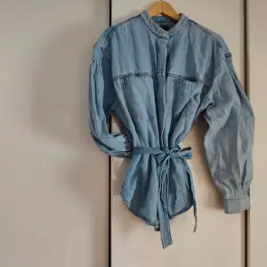 Svinsnygg jeansskjorta/jacka med farfarskrage, tryckknappar, knytning i midjan och ballongärm. Knappt använd. Storl. L från Lindex