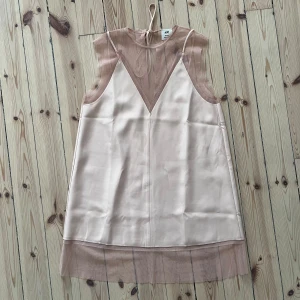 Beige ärmlös klänning från H&M - En elegant beigerosa ärmlös klänning från H&M studio. Använd en gång. Klänningen har en transparent övre del och en satinliknande mittendel och sen en transparent nederdel. Den har en rak passform. väldigt söt! barbie! har dock en fläck som troligtvis går bort i tvätt