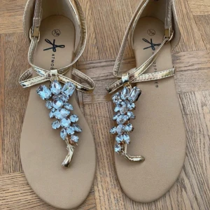 Guldiga sandaler med strassdetaljer från ATM - Eleganta guldiga sandaler från Atmosphere i storlek 38. Sandalerna har en T-rem design med stora, glittrande strassdetaljer på remmen. De har ett justerbart spänne för en bekväm passform.