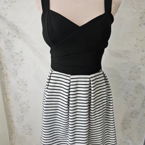 Svart och vit randig klänning från Sandra Fellini - En elegant klänning från Sandra Fellini med en svart överdel och en vit och svart randig kjol. Klänningen har breda axelband och en dragkedja i ryggen. Den är tillverkad i Italien. Slits på sidan.