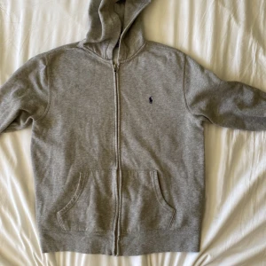  Ralph Lauren zip  - En grå huvtröja från Polo Ralph Lauren med dragkedja framtill. Tröjan har en broderad logotyp på vänster bröst och två framfickor. Den är tillverkad av bomull och har långa ärmar. Ny pris 1000 mitt 449 skick 8/10