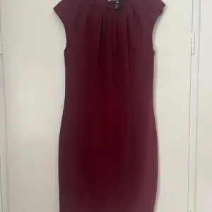Vinröd klänning som går ner till mitten på låret. Dragkedja och knapp i ryggen