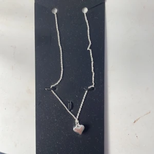 Silverfärgat halsband med hjärthänge - Ett elegant silverfärgat halsband med ett hjärthänge. Halsbandet har en tunn kedja och ett litet hjärta som hänge.