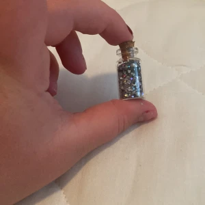 Glittrig ögonskugga i flaska - En liten flaska med glittrig ögonskugga i silver och flerfärgade nyanser. Flaskan har en kork och är lätt att använda för att applicera glittret på ögonlocken.