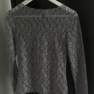 Grå spetsblus - En elegant grå spetsblus med långärmad design. Blusen har ett vackert blommönster och är tillverkad av spetsmaterial.