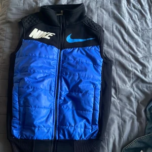 Blå och svart dunväst från Nike - En blå och svart dunväst från Nike med dragkedja framtill. Västen har en hög krage och är dekorerad med Nike-logotypen i vitt och blått på framsidan samt en stor vit Nike-logotyp på baksidan.