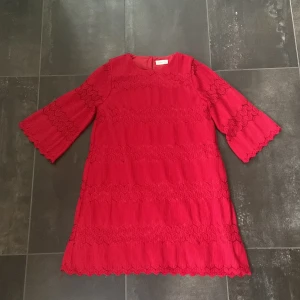 Mini klänning - En röd klänning från Name It med spetsdetaljer. Klänningen har en rund hals och är dekorerad med horisontella spetsband. Den har en knappstängning i ryggen och är fodrad för extra komfort., är strl 146 men är ganska stor i strl som kan passa även xxs/ xs, perfekt klämning inför julen, köpt för 500kr säljer för 180kr, 
