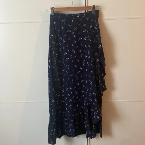 Blommig kjol från Neo Noir - En mörkblå lång omlottkjol med små blommor i rosa och vitt från Neo Noir. Kjolen har volangdetaljer och ett knytband i midjan.