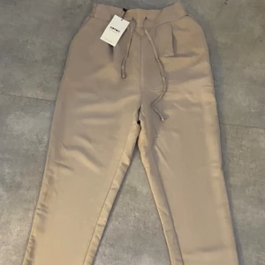 Tygbyxor- beige - Beiga kostymbyxor med knytband och raka ben från koton. De har även resår i baksidan av midjan (syns inte på bild). Aldrig använda och med prislapp säljer då de inte kommer till användning.