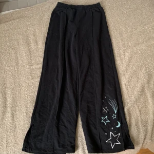 Svarta byxor med stjärnmönster från H&M - Ett par svarta byxor från H&M med ett tryck av stjärnor, måne och komet i blått och vitt på ena benet. Byxorna har en elastisk midja och är vida i modellen. Dem har en slits nere vid benen.