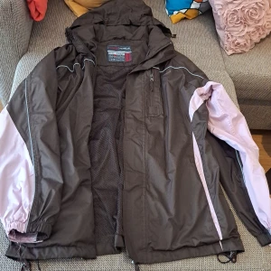 Svart och rosa vindjacka från Karrimor - En svart vindjacka och regnjacka från Karrimor med rosa detaljer på ärmarna. Jackan har en dragkedja framtill, en bröstficka med dragkedja och justerbara ärmslut med kardborreband. Den har även en huva och meshfoder på insidan för bättre ventilation.