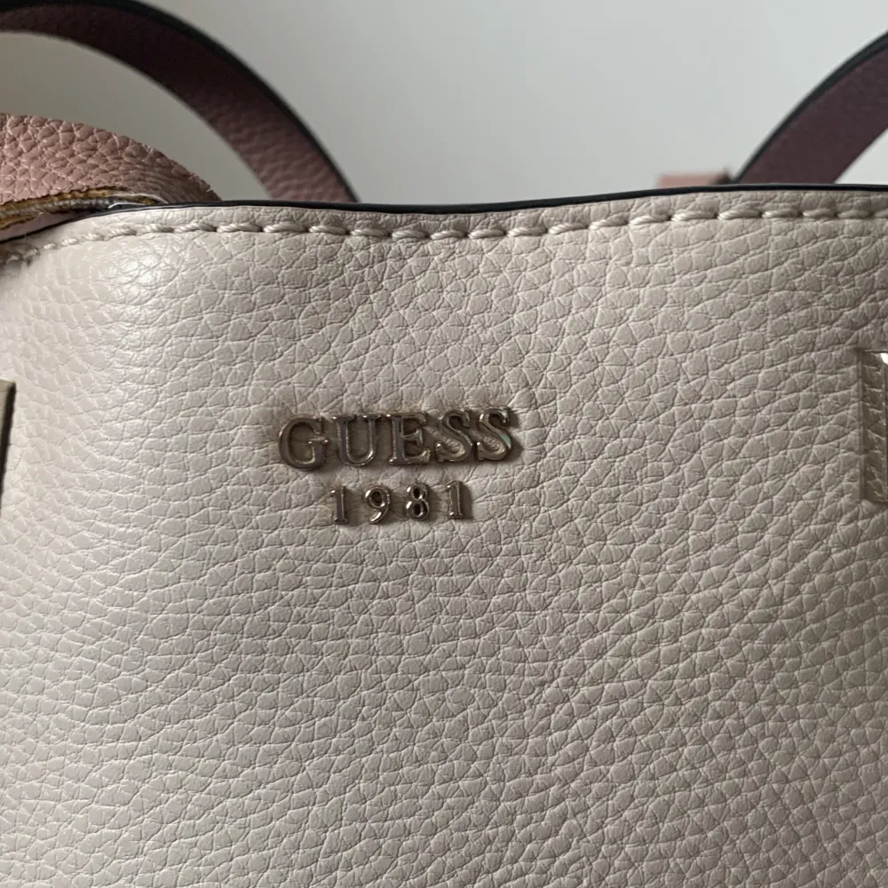 En  handväska från Guess i beige brun färg. Väskan har två handtag och en rymlig insida med flera fack. Den är tillverkad i skinn och har Guess-logotypen. Den är äkta och nypris låg runt två tre tusen. Lite skadad på vissa ställen därav priset.. Laukut & Käsilaukut.