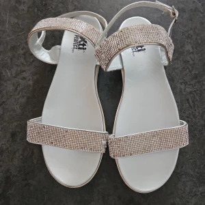 Silvriga sandaler med strass  - Silvriga sandaler med strass. Sandalen har en platt sula och en rem runt foten. Sandalen är i bra skick. Men finns defekter