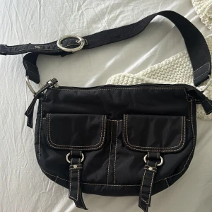 Svart axelväska med fickor och spännen - En svart axelväska med justerbar rem och silverfärgade detaljer. Väskan har två framfickor med spännen och en dragkedjeficka på baksidan. Insidan är rymlig med en extra dragkedjeficka. Perfekt att ha på tex klubb när man vill inte ha en för stor väska :)