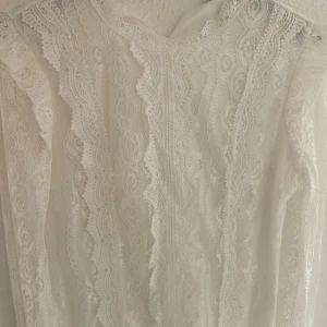 Blus  - En elegant vit spet blus med detaljerade spetsmönster och långärmad design. blusen har en hög hals och är tillverkad av ett genomskinligt spetsmaterial som ger en romantisk och sofistikerad look.