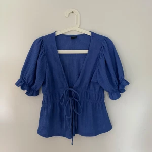 Blå blus med puffärmar - En blå blus med puffärmar och knytning framtill. Blusen har en djup v-ringning och elastisk midja för en bekväm passform.