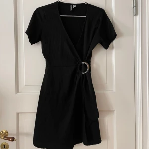 Omlottklänning - Svart klänning från H&M i storlek 40 men skulle säga att den är liten i storlek och passar 36-38 mer. Omlott och stängs med ”skärpknäppe” (se bild). Struktur på materialet men stretchig. Stretchigt material. Aldrig använd, i nyskick. 