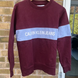 Tjocktröja - Säljer denna något urtvättade tjocktröja från Calvin Klein som är vinröd med ett ljusblått sträck över bröstet och vänster arm på framsidan. Den är i använt skick.