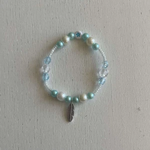 Egengjort pärlarmband  - Ett egengjort pärlarmband med färgerna blå och vit i olika nyanser med en blandning pärlsorter. Armbandet har en berlock i silver som avbildar en fjäder. 