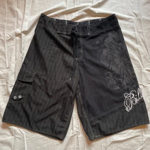 Sommarshorts - Svarta Billabong shorts att ha på sig hela sommaren. Passar på stan, stranden och i stugan! Dödskalle på vänstra sidan, ficka på höger sida. Kardborre och snörning som knäppe. Två små hål innanför kardborren (se bild 3) som ej syns när de är på.  