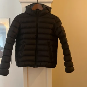 Colmar dunjacka - Svart dunjacka med luva och fickor och grått foder. Storlek 12Y eller 154 cm. I bra skick, sparsamt använd. Ordinarie pris 3299 sek. 