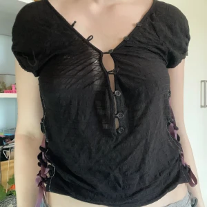 Svart topp korsettsnörning - En svart topp med korsettsnörning med lila band på båda sidor. Supergullig i ett tunt tyg som faller jättefint. Från Filippa k från början.