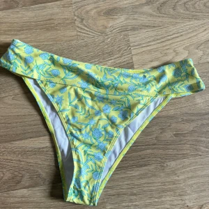 Gul blommig bikiniunderdel - En gul bikiniunderdel med blommigt mönster i blått. Underdelen har en bred linning. liten i storlek. Helt ny